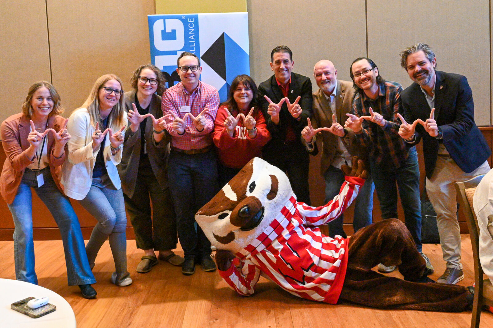 A group of people posing for a photo make the letter W with their fingers. Bucky Badger lays in front of the group.